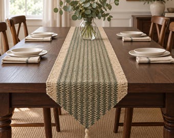 May include: A rectangular woven table runner featuring a central dark green and tan striped pattern with natural tan borders. The runner has a textured finish and a single decorative tassel hanging from the pointed end, centered on a dark wood table.