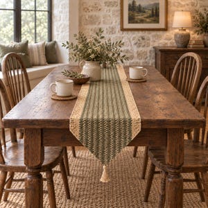 May include: A rectangular table runner featuring a woven green and beige textured pattern with a central olive green stripe, finished with a small beige tassel at the pointed end, displayed on a dark brown wooden dining table with two mugs.
