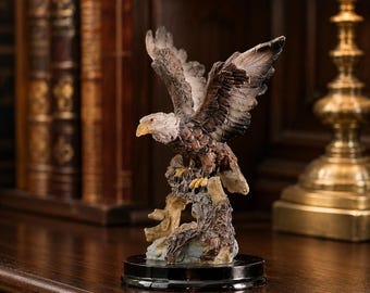 May include: A bald eagle figurine with outstretched wings, featuring a white head, brown body, and yellow beak, perched on a textured tree branch base. The sculpture is mounted on a circular black stand, showcasing detailed feather textures.