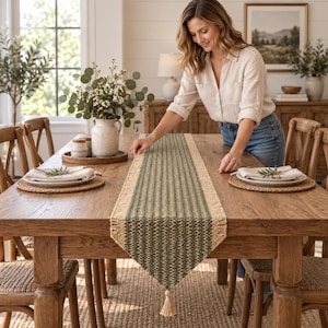 May include: A rectangular table runner featuring a woven texture with a central dark green stripe flanked by natural tan borders. The runner is finished with a single decorative tassel at the pointed end, showcasing a detailed, textured weave pattern.