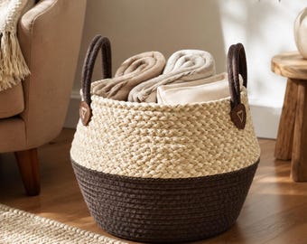May include: A round storage basket featuring a two-tone design with a natural light beige woven upper section and a dark brown woven lower section. The basket includes two dark brown handles attached with triangular brown leather-like patches.