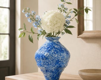 May include: A textured, bulbous vase with a mottled blue and white finish, containing a bouquet of white hydrangea flowers and tall, slender stems with small light blue blossoms, creating a vibrant contrast against the neutral surroundings.