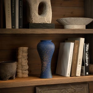 Peut inclure: Un vase en céramique bleue avec une finition texturée et tachetée posé sur une étagère en bois. Le vase présente un col étroit et une base plus large, placé entre un grand objet beige texturé en forme de pilier et des livres aux dos clairs.