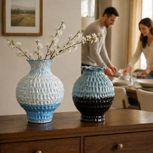 Pode incluir: Dois vasos de cerâmica com superfície texturizada e bordas onduladas. O vaso maior apresenta um degradê em branco e azul claro, enquanto o menor tem um degradê em azul e preto. O vaso maior contém um ramo com flores brancas.