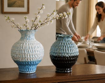 May include: Two ceramic vases with a textured, dimpled surface and ruffled rims. The larger vase features a white and light blue gradient, while the smaller vase has a blue and black gradient. The larger vase holds a branch with white blossoms.