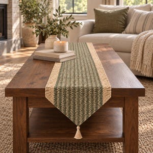 May include: A rectangular table runner featuring a central panel of woven green and white textured fabric, bordered by natural tan jute-like material on both sides. The runner ends in a pointed tip finished with a single small cream-colored tassel.
