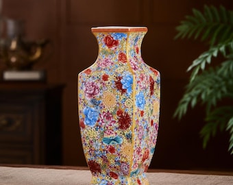 May include: A square-shaped ceramic vase featuring a vibrant yellow background densely decorated with a colorful floral pattern. The design includes various blossoms in shades of red, blue, pink, and white, covering the entire exterior surface.