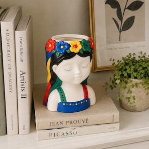 Op de afbeelding: Een keramische plantenbak in de vorm van een hoofd met gesloten ogen, voorzien van een kleurrijke bloemenkrans in rood, blauw, geel en groen. Het figuur heeft zwart haar en draagt een blauwe top met groene en rode bandjes met stippen.