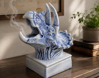 May include: A ceramic triceratops head incense burner featuring a blue and white glazed finish with a textured, scale-like surface. The dinosaur head is mounted on a square base, with thin wisps of smoke rising from its open, roaring mouth.