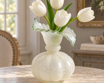May include: A translucent white glass vase with a rounded, ribbed body and a wide, ruffled rim. The vase contains four white tulips with green stems and leaves. The glass has a subtle, swirled texture throughout its surface and a clear base.