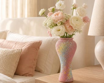 May include: A tall, slender vase with a textured, speckled finish in shades of pink, yellow, and blue, holding a bouquet of white, cream, and soft pink flowers including ranunculus and small filler blossoms with green stems.