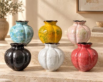 May include: A collection of six small, round, pumpkin-shaped ceramic vases with ruffled rims. The vases feature glossy, mottled finishes in blue, yellow, pink, black, white, and red, each with a dark brown interior rim and base.