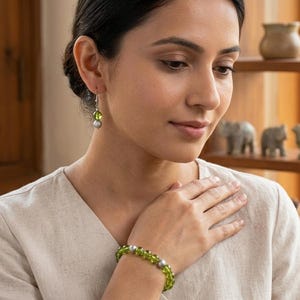 Puede incluir: Un conjunto de joyería compuesto por una pulsera y un pendiente individual con cuentas de color verde lima translúcido y cuentas esféricas de tono plateado opaco. La pulsera alterna cuentas verdes y plateadas, el pendiente tiene un gancho plateado.