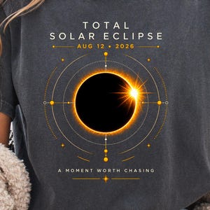 May include: A charcoal gray short-sleeve t-shirt featuring a graphic of a total solar eclipse with the text TOTAL SOLAR ECLIPSE AUG 12 2026 and A MOMENT WORTH CHASING. The design includes golden orbital lines and a bright sun flare effect.