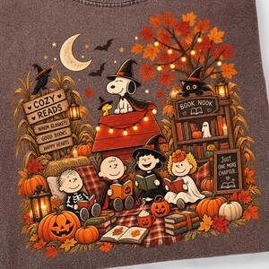 May include: A brown crewneck t-shirt featuring a graphic of Peanuts characters in Halloween costumes reading books. The design includes a doghouse, pumpkins, autumn leaves, and text reading COZY READS, BOOK NOOK, and JUST ONE MORE CHAPTER.
