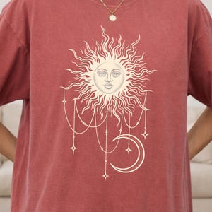 May include: A brick red short-sleeve t-shirt featuring a cream-colored graphic print on the chest. The design depicts a sun with a human face surrounded by radiating rays, with dangling chains, stars, and a crescent moon hanging below.