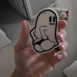 May include: A white, die-cut acrylic or plastic ornament featuring a black-outlined cartoon ghost character. The ghost is depicted with its hands on its hips, wearing a black thong, and has a small hole at the top for hanging purposes.