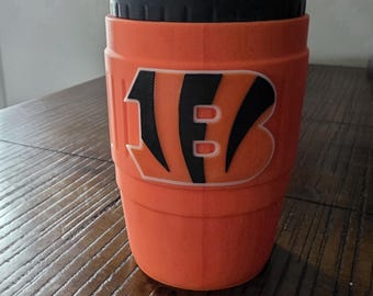 May include: An orange insulated beverage cup featuring a black lid and the Cincinnati Bengals logo in black and white on the front. The cup has a textured, ribbed exterior surface and a cylindrical shape designed to hold a standard drink container.