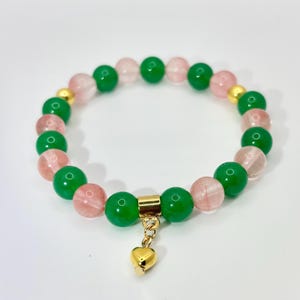 May include: A beaded bracelet featuring alternating round green and translucent pink beads. The bracelet includes small gold-toned spherical spacers and a central gold-toned cylindrical bead from which a small gold-toned heart charm hangs.