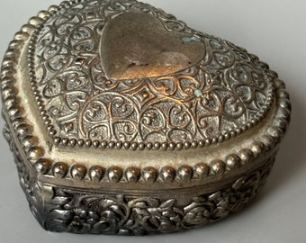 Puede incluir: Una caja de corazón de plata con un diseño ornamentado. La caja tiene un corazón en relieve en el centro y un borde de cuentas.