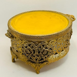 May include: A gold-tone metal trinket box with a round, mirrored top and a decorative base with floral accents. The box has a filigree design and sits on four feet.