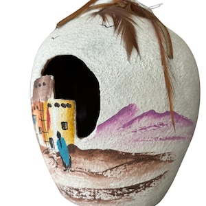 Puede incluir: Maceta de cerámica blanca con una escena pintada de un paisaje desértico con un edificio de estilo pueblo y montañas moradas. La maceta tiene una pluma marrón en la parte superior.
