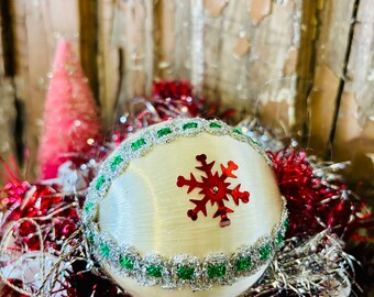 May include: A white Christmas ornament with a red snowflake design and a green and silver trim. The ornament is surrounded by red and silver tinsel.