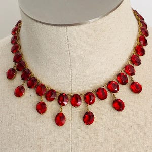May include: A vintage necklace featuring large, faceted red glass stones. The stones are oval-shaped and dangle from a gold-toned chain. The necklace is displayed on a mannequin, showcasing its design and color.