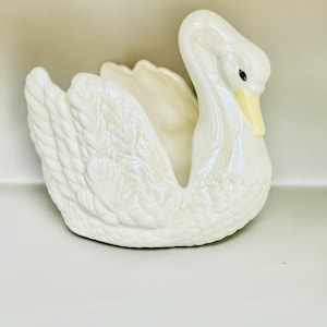 May include: A white ceramic swan planter with iridescent detailing. The swan has a yellow beak and black eyes. The planter is designed to hold small plants or flowers.