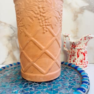 May include: A terracotta-colored cylindrical vase with embossed grape and leaf designs, set on a mosaic tray with blue and turquoise tiles. A small white pitcher with red splatter paint is also visible.