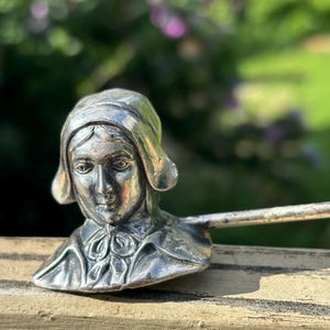 Puede incluir: Una escultura de metal plateado de un busto de mujer, que lleva un gorro y un cuello con volantes. La escultura está montada sobre una varilla de metal, posiblemente un utensilio para servir o una pieza decorativa. El fondo está borroso, con follaje verde y toques de flores moradas.