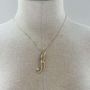 May include: A gold-colored necklace with a pendant in the shape of the letter "K". The pendant has a decorative design with small, clear stones on the left side. The necklace is displayed on a mannequin.