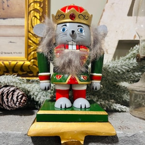 Puede incluir: Un soporte de calcetín navideño de madera con forma de cascanueces de ratón. El ratón es gris con una corona roja y dorada, uniforme verde y rojo y barba de piel sintética. La base es verde y dorada.