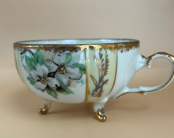 May include: A white porcelain teacup with a floral design and gold accents. The cup has three legs and a heart-shaped handle.