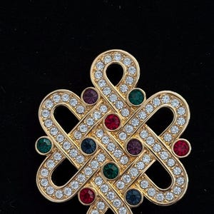 May include: Gold-toned brooch with a repeating knot design, encrusted with clear rhinestones. The brooch features small, colorful gemstones in red, green, purple, and blue. The brooch is set against a black background.