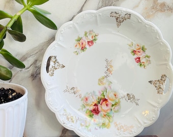 May include: White, scalloped-edge serving platter with a floral rose design in shades of pink, yellow, and red. The platter has gold accents and two handles. A potted plant is in the background.