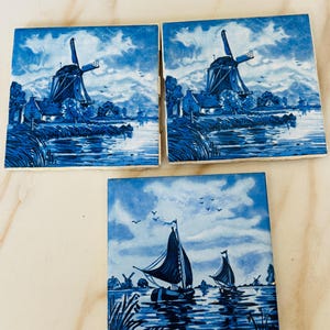 May include: Three square ceramic tiles with blue and white Dutch windmill and sailboat scenes. The tiles feature a classic Delftware design with windmills, boats, water, and sky. The tiles are arranged on a light-colored surface.