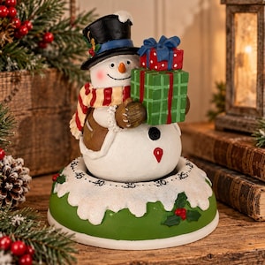 May include: A snowman figurine wearing a black top hat and a red and white striped scarf, holding a red gift box and a green gift box. The snowman sits on a circular kitchen timer base with a green bottom and a white snow-covered top edge.