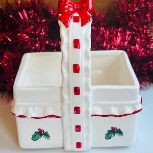 Puede incluir: Una cesta de cerámica blanca con un asa de lazo rojo. La cesta tiene forma rectangular con borde con volantes y está decorada con detalles rojos y hojas de acebo. Una decoración festiva.