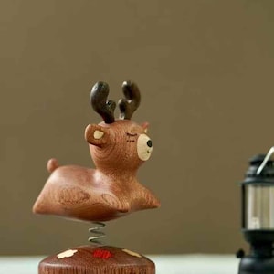 May include: A wooden deer figurine with dark brown antlers and a light-colored muzzle, featuring closed eyes and a small tail. The deer is mounted on a metal spring attached to a round wooden base decorated with small white and red floral accents.