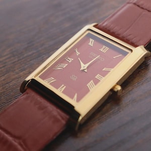 May include: A rectangular wristwatch with a gold-tone case and a deep red dial featuring gold Roman numeral hour markers. The watch has gold-tone hands and a textured brown leather strap with a visible grain pattern and a side adjustment crown.