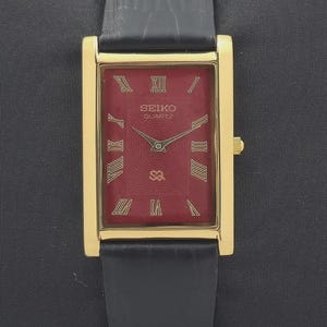May include: A rectangular Seiko Quartz wristwatch featuring a gold-tone case and a deep red dial with gold Roman numeral hour markers. The watch has a black leather strap with a textured finish and gold-coloured hands for hours and minutes.