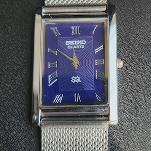 May include: A rectangular Seiko Quartz wristwatch featuring a deep blue dial with a concentric circular pattern, silver-tone Roman numeral hour markers, and gold-tone hands. The watch has a polished silver-tone metal case and a silver mesh strap.