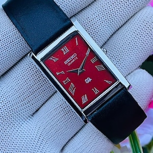 Op de afbeelding: Een rechthoekig Seiko Quartz polshorloge met een felrode wijzerplaat, zilverkleurige Romeinse cijfers als uurmarkeringen en bijpassende zilveren wijzers. Het horloge heeft een gepolijste zilverkleurige metalen kast en een gladde zwarte leren band.