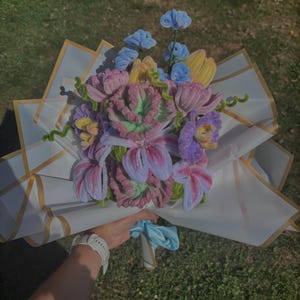 Può includere: Un bouquet di fiori realizzati a mano con scovolini in tonalità rosa, viola, giallo e blu, con petali testurizzati e steli verdi. La composizione è avvolta in carta bianca con bordi dorati e legata con un nastro azzurro alla base.