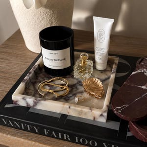 Könnte beinhalten: Ein quadratisches Marmortablett mit violetten und weißen Adern enthält eine schwarze Byredo Bibliotheque Kerze, eine weiße Dieux Handcremetube, einen transparenten Glasparfümflakon, eine goldene muschelförmige Schale, ein goldenes Armband und einen Ring.