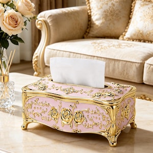 May include: A rectangular tissue box holder featuring a light pink finish with ornate gold-toned metal accents. The design includes raised floral motifs, scrolling vine patterns, and small embedded clear crystals along the top edge and four feet.