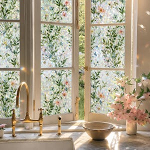 Könnte beinhalten: Eine Fensterfolie mit einem botanischen Muster aus grünen Blättern sowie kleinen rosa und blauen Blüten, die eine prismatische, geometrische Textur aufweist, welche das Licht in Regenbogenfarben bricht und auf den Glasscheiben eines Küchenfensters klebt.