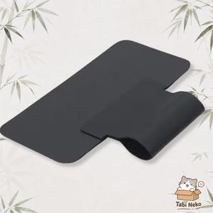May include: A matte black, rectangular silicone desk mat featuring an integrated, curved cable management loop on one side. The surface is smooth and solid black, designed to organize cords while providing a flat workspace area for various items.