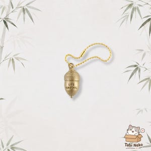 Può includere: Un ciondolo a forma di ghianda in metallo color oro attaccato a una catenella a palline abbinata. La ghianda presenta una calotta testurizzata e una base liscia con la scritta LIFE GROWS. incisa in lettere nere sulla superficie anteriore.